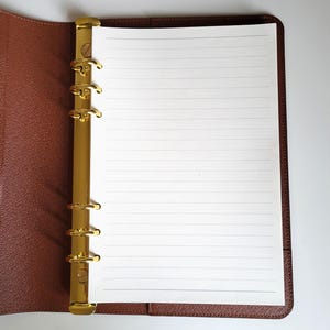 May include: A brown textured leather binder featuring a gold-colored six-ring metal mechanism. The binder is open, displaying a single sheet of white lined paper held in place by the rings. The interior cover is lined with matching brown material.