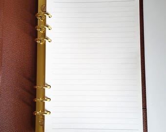 May include: A brown textured leather binder featuring a gold-colored six-ring metal mechanism. The binder is open, displaying a single sheet of white lined paper held in place by the rings. The interior cover is lined with matching brown material.