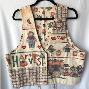 May include: A sleeveless waistcoat featuring a cream background with a harvest-themed print including scarecrows, pumpkins, and autumn leaves. The front displays the text "HARVEST" in green letters, a small crescent moon, and a wheelbarrow labelled "The Pumpkin Patch."
