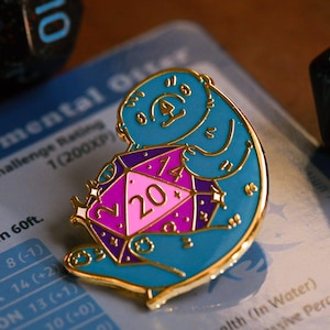 May include: A gold-tone enamel pin featuring a blue otter holding a purple and pink 20-sided die. The die has a gold border and a white star on each side. The otter is holding the die with its paws and has a white star on its chest. The pin is on a table with a character sheet in the background.