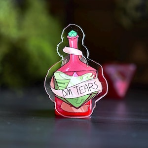 May include: A clear acrylic pin shaped like a potion bottle. The bottle is red with a green liquid inside and a banner that reads "DM TEARS". A green gem tops the bottle.