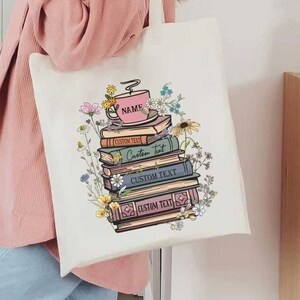 May include: A white tote bag with a design of a stack of books with a coffee cup on top, surrounded by flowers. The text on the bag reads "NAME", "CUSTOM TEXT", "CUSTOM TEXT", "CUSTOM TEXT", and "CUSTOM TEXT".