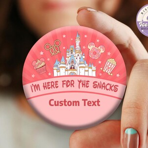 May include: A round, pink button with the text "I'M HERE FOR THE SNACKS" and "Custom Text". The button features illustrations of a castle, snacks, and stars. The button is held by a person.