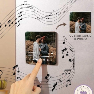 May include: A custom music and photo magnet with a black and white music note design. The magnet features a photo of a couple in a forest setting. The text "Song Name" and "Artist Name" are visible on the magnet. The magnet is on a silver refrigerator.
