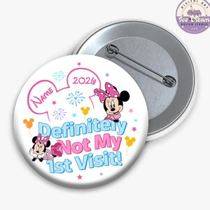 May include: A white button with a Minnie Mouse design and the text "Definitely Not My 1st Visit!" in blue and pink. The button features a pink bow, stars, and the year 2026. A second button is visible in the background.