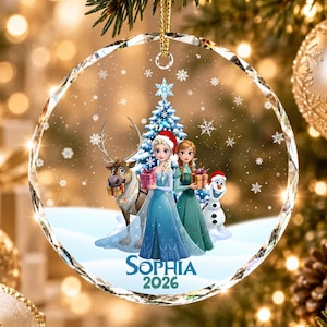 May include: A round, clear acrylic Christmas ornament featuring a printed illustration of Elsa, Anna, Sven, and Olaf from Frozen. The characters stand before a blue Christmas tree with snowflakes. The name Sophia and the year 2026 are printed below.