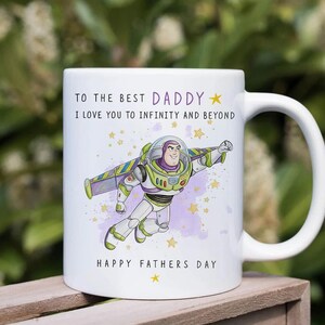 Puede incluir: Taza de cer&aacute;mica blanca con una ilustraci&oacute;n de dibujos animados de Buzz Lightyear volando, con el texto "TO THE BEST DADDY" y "I LOVE YOU TO INFINITY AND BEYOND". La taza tambi&eacute;n incluye "HAPPY FATHERS DAY" y estrellas.