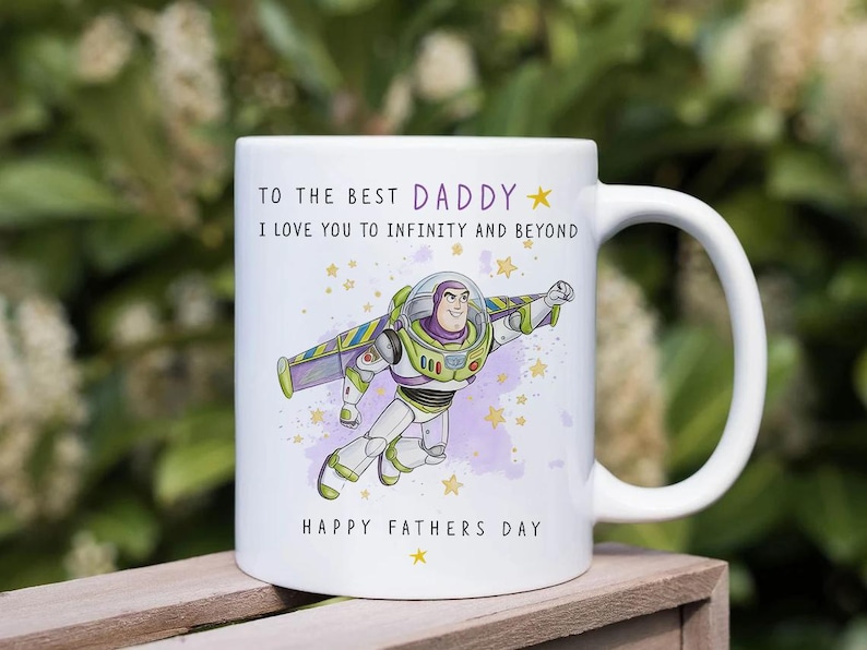 Puede incluir: Taza de cer&aacute;mica blanca con una ilustraci&oacute;n de dibujos animados de Buzz Lightyear volando, con el texto "TO THE BEST DADDY" y "I LOVE YOU TO INFINITY AND BEYOND". La taza tambi&eacute;n incluye "HAPPY FATHERS DAY" y estrellas.