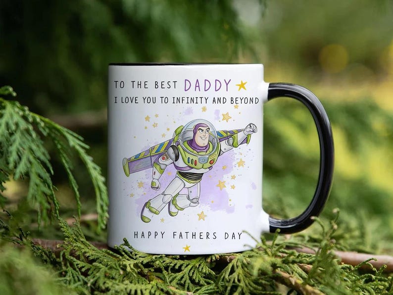 Puede incluir: Taza de cer&aacute;mica blanca con asa y borde negros, con una imagen de dibujos animados de Buzz Lightyear y el texto "TO THE BEST DADDY, I LOVE YOU TO INFINITY AND BEYOND, HAPPY FATHERS DAY". La taza est&aacute; decorada con estrellas.
