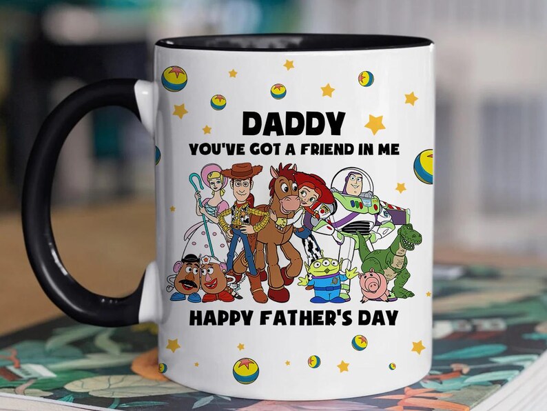 Puede incluir: Taza de cer&aacute;mica blanca con interior y asa negros. La taza presenta un gr&aacute;fico colorido de personajes de la pel&iacute;cula Toy Story, con el texto "DADDY YOU'VE GOT A FRIEND IN ME HAPPY FATHER'S DAY". Estrellas amarillas y gr&aacute;ficos de bolas de Toy Story est&aacute;n dispersos alrededor de la taza.