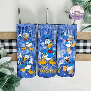 May include: Three blue and white tumbler cups with a glitter finish. Each cup features a different image of Donald Duck in a sailor outfit. The middle cup has the name "Michael" written in gold lettering.