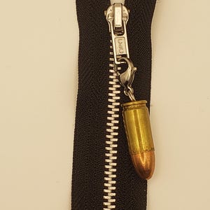 May include: Close-up of a black zipper with a silver pull and a bullet-shaped charm. The bullet charm has a brass casing and a copper-colored tip, adding a unique detail to the zipper.