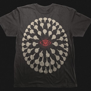 May include: Dark gray t-shirt featuring a circular design of white teardrop shapes surrounding a red eye. The graphic is centered on the front of the shirt. The shirt is made of a soft material.