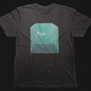 Puede incluir: Camiseta gris oscuro con un gran gráfico turquesa de la tecla "Esc" de un ordenador. La tecla tiene un aspecto texturizado y la palabra "Esc" está impresa en blanco. Es una prenda de vestir informal.