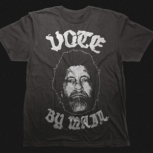 Op de afbeelding: Donkergrijs t-shirt met een witte afbeelding. De afbeelding toont het woord "VOTE" boven een portret van een man met een baard en krullend haar, en de woorden "BY MAIL" eronder. Het shirt is gemaakt van een zacht materiaal.