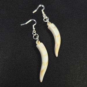 May include: A pair of earrings featuring curved, ivory-colored pendants with brown marbling. The pendants are suspended from silver-colored fish hook ear wires. The earrings are displayed against a dark background.
