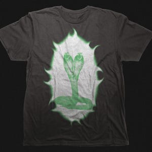 May include: Dark grey t-shirt with a green cobra design. The cobra is centrally placed, with a white and green outline. The design has a vintage, slightly distressed appearance. The t-shirt is made of a soft material.
