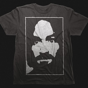 Könnte beinhalten: Dunkelgraues T-Shirt mit einer weißen Grafik eines Gesichts in einem weißen rechteckigen Rahmen. Das Gesicht hat einen Schnurrbart und einen Bart. Das T-Shirt ist aus weichem Material.