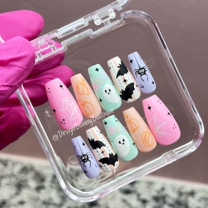 May include: A set of ten press-on nails in a clear plastic case, featuring pastel pink, peach, mint green, and lavender colors. Designs include white spiderwebs, orange pumpkins, black bats, a white ghost with the text BOO, and black spiders.