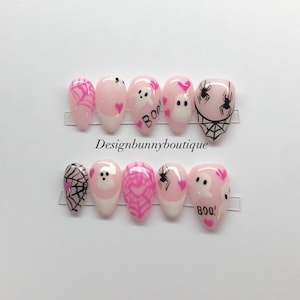 May include: Set of pink and white Halloween-themed false nails. Designs include spiderwebs, ghosts, hearts, and the word "BOO!" in black. The nails are arranged on clear plastic holders, with the text "Designbunnyboutique" visible.