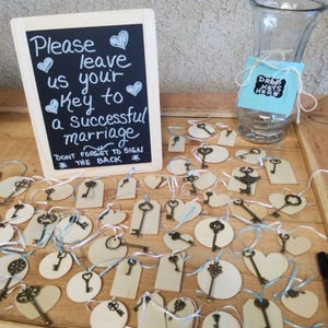 May include: A wedding guest book alternative featuring a chalkboard sign with the text "Please leave us your Key to a successful marriage." Wooden tags and heart-shaped cutouts with antique-style keys are arranged on a wooden surface.
