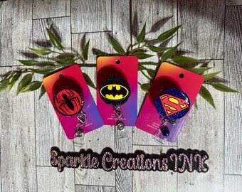 May include: Three superhero themed retractable badge holders. The holders are red with a black glitter background. The first holder features a red and black spider-man logo, the second holder features a black and yellow batman logo, and the third holder features a red and blue superman logo.