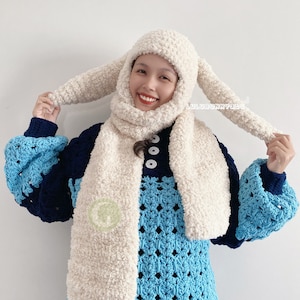 May include: A white crocheted hat with long bunny ears and a matching scarf. The hat and scarf are made of a fluffy yarn. The person wearing the hat and scarf is smiling.