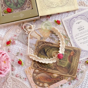 Può includere: Una collana di pizzo bianco con un ciondolo a forma di fiore rosso. La collana è su un tavolo con diverse carte dei tarocchi, tra cui "L'alveare d'oro" e "Il fascino del burlone".