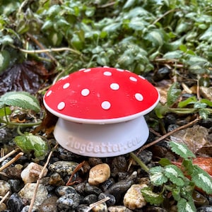 Könnte beinhalten: Ein rotes, pilzförmiges Gartenzierobjekt mit weißen Punkten. Die Pilzkappe sitzt auf einem weißen Sockel, der auf kleinen Steinen und Grün liegt. Der Text "SlugGetter.com" ist auf der Basis aufgedruckt.