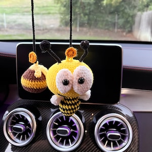 May include: A crocheted bee car charm in yellow and black, with large white-rimmed eyes and black antennae. A smaller crocheted honey pot charm hangs nearby. The bee has a black and yellow striped abdomen.