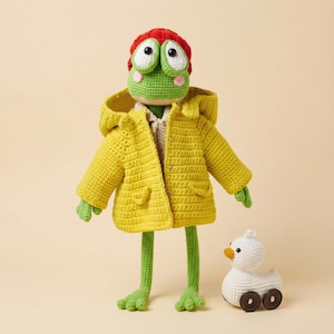 Könnte beinhalten: Eine handgefertigte Häkelfroschpuppe mit gelber Kapuzenjacke und roter Mütze. Der Frosch hat große, ausdrucksstarke Augen und grüne Gliedmaßen. Eine weiße Häkelente mit braunen Rädern sitzt in der Nähe. Die Puppe ist ein skurriles, handgefertigtes Objekt.