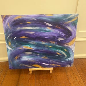 Puede incluir: Pintura abstracta sobre lienzo con trazos en espiral de pintura p&uacute;rpura, turquesa, dorada y blanca. La pintura se muestra en un peque&ntilde;o caballete de madera.