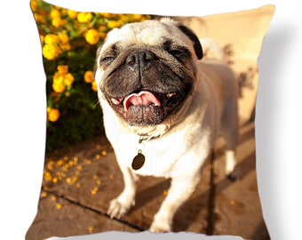 May include: A close-up of a pug dog with its tongue sticking out, looking up at the camera. The dog is standing on a wooden surface with yellow flowers in the background.