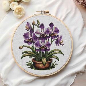 Puede incluir: Un patrón de punto de cruz con una flor de orquídea púrpura y blanca en un aro de bordado de madera. El aro está sobre un fondo de tela blanca.