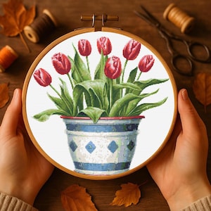 May include: A cross-stitch embroidery hoop featuring a vibrant floral design. The artwork showcases red tulips in a blue and white patterned pot. The hoop is held by hands, with a wooden background and craft supplies visible.