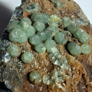 May include: A close-up of a rock with a cluster of small, green, round crystals. The crystals are clustered together in a dense formation, and they have a slightly translucent appearance. The rock is a dark brown color, and it has a rough, textured surface.
