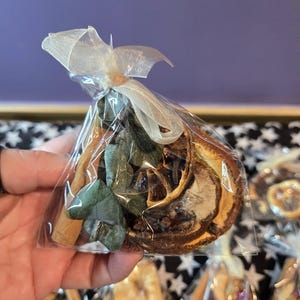 Puede incluir: Una bolsa transparente de celofán atada con una cinta blanca, llena de rodajas de naranja secas, palitos de canela y hojas verdes. La bolsa se sostiene en una mano, con otras bolsas similares visibles en el fondo.