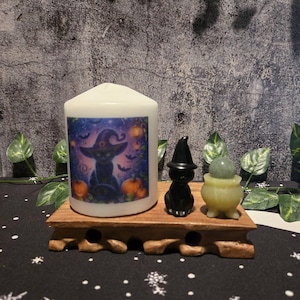 Peut inclure: Une bougie pilier blanche ornée d'un motif représentant un chat noir portant un chapeau de sorcière entouré de citrouilles et de chauves-souris. À côté se trouvent une figurine de chat noir et un chaudron vert clair avec une pierre ronde.