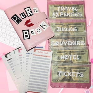 May include: A pink budget binder with the text "Burn Book" and a red lip graphic. Clear pink pouches labeled "Travel Expenses," "Dining," "Souvenirs," "Hotel," and "Tickets" are filled with cash. Budget sheets and a pink pen are also visible.