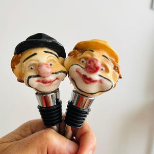 May include: Two decorative wine bottle stoppers featuring clown faces. One clown wears a black hat, the other a yellow hat. Both have painted facial features, including red noses and smiling mouths. The stoppers have silver metal stems and black rubber seals.