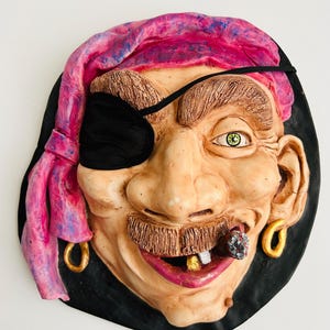 May include: A sculpted pirate face with a pink bandana, black eye patch, and gold hoop earrings. The pirate has a brown mustache, a cigar in his mouth, and a gold tooth. The face is set against a black background.