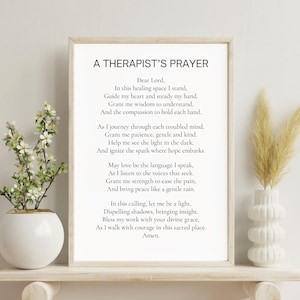 May include: A framed print with the text "A Therapist's Prayer" in black font. The print is displayed on a white shelf with a white vase holding dried flowers on the right and a white vase with a plant on the left.