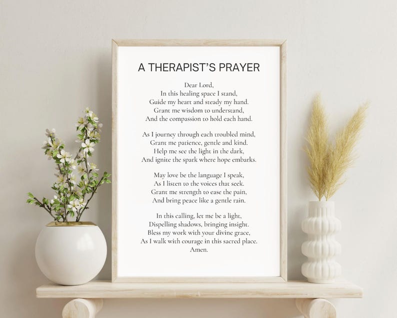 May include: A framed print with the text "A Therapist's Prayer" in black font. The print is displayed on a white shelf with a white vase holding dried flowers on the right and a white vase with a plant on the left.