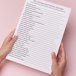 May include: A white sheet of paper titled "IDENTIFYING EMOTIONAL TRIGGERS" is held by two hands against a pink background. The paper contains a list of prompts to help identify feelings and experiences.
