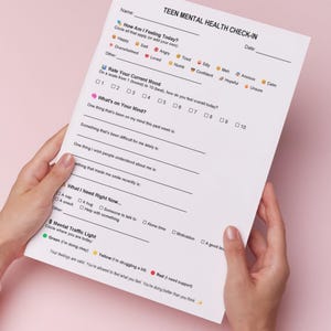 Puede incluir: Una persona sostiene un papel blanco titulado "TEEN MENTAL HEALTH CHECK-IN". El formulario incluye secciones para calificar el estado de &aacute;nimo, identificar pensamientos y necesidades. El fondo es rosa.