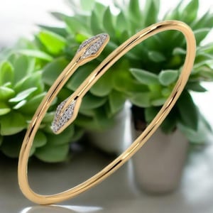 May include: A gold-coloured bangle bracelet with two decorative ends. Each end features a leaf-like design encrusted with small, sparkling crystals. The bracelet is set against a backdrop of green plants and white pots.