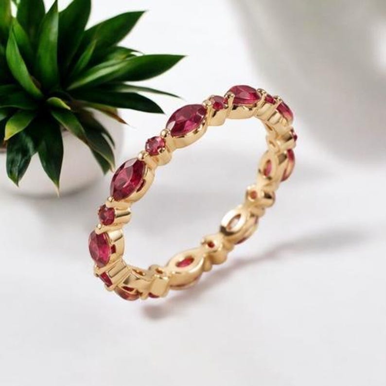 May include: A gold band ring featuring an alternating pattern of marquise-cut and round-cut red gemstones set along the entire circumference. The metal has a polished finish, and the stones are secured in individual gold settings.