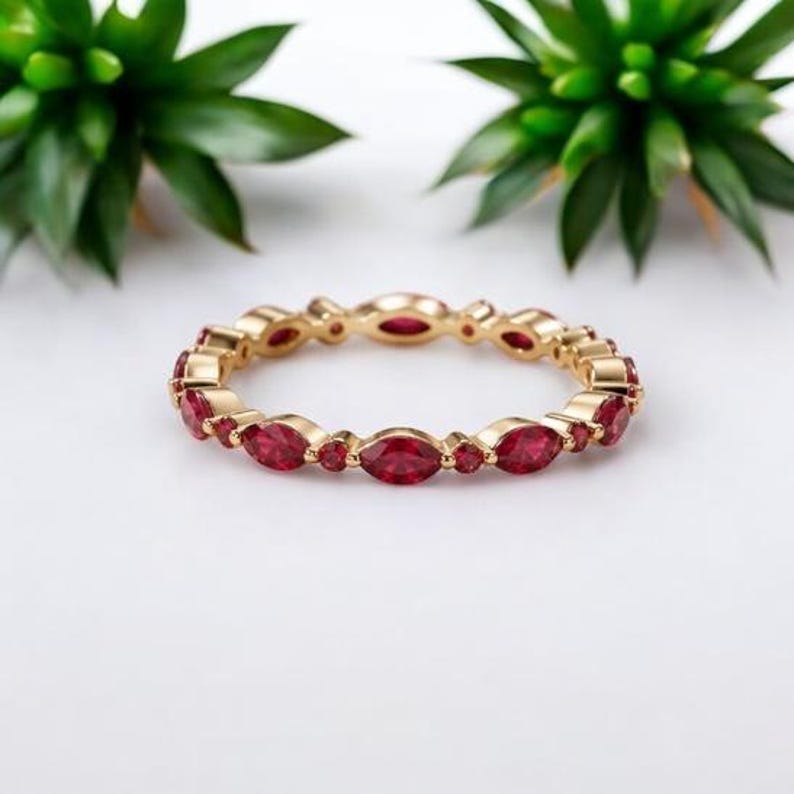 May include: A gold band ring featuring an alternating pattern of marquise-cut and round-cut red gemstones. The ring is constructed with a polished metal finish, showcasing a continuous design of faceted stones set securely within the gold frame.