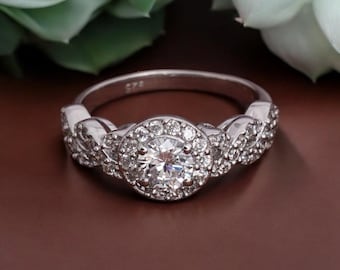 May include: A silver ring with a large, round, clear center stone surrounded by smaller, clear stones. The band features a braided design on either side of the center stone. The ring is set against a brown background.