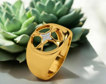 Puede incluir: Anillo dorado con un diseño geométrico que presenta un patrón en forma de estrella en el centro, incrustado con pequeñas piedras brillantes. El anillo tiene una banda gruesa y un acabado pulido, sobre un fondo de plantas suculentas verdes.